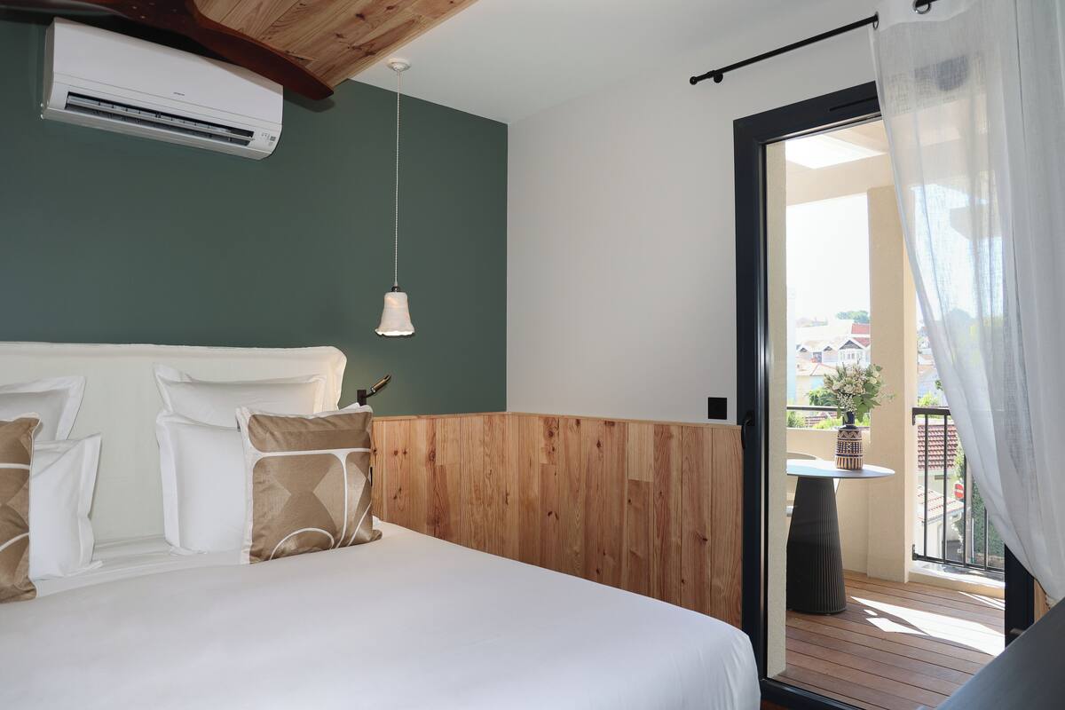 A bedroom with a wooden headboard, a bed with white bedding, a hanging pendant light, and a view of a balcony with a table and potted plant.