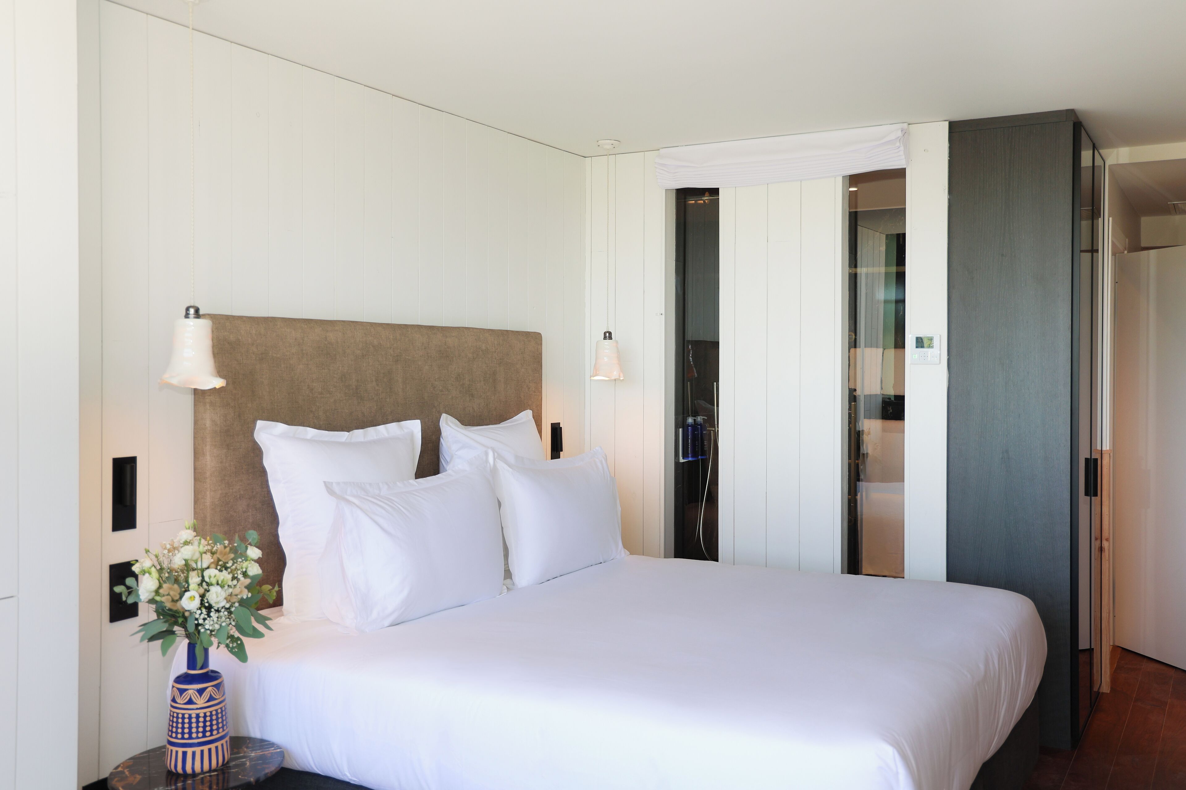Habitación de hotel con una cama grande, ropa de cama blanca, un florero con flores y un espejo en la pared.