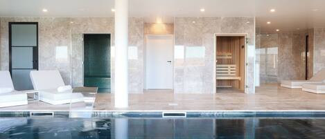 A modern indoor pool area with clear blue water, white lounge chairs, and a spa room with wooden benches.