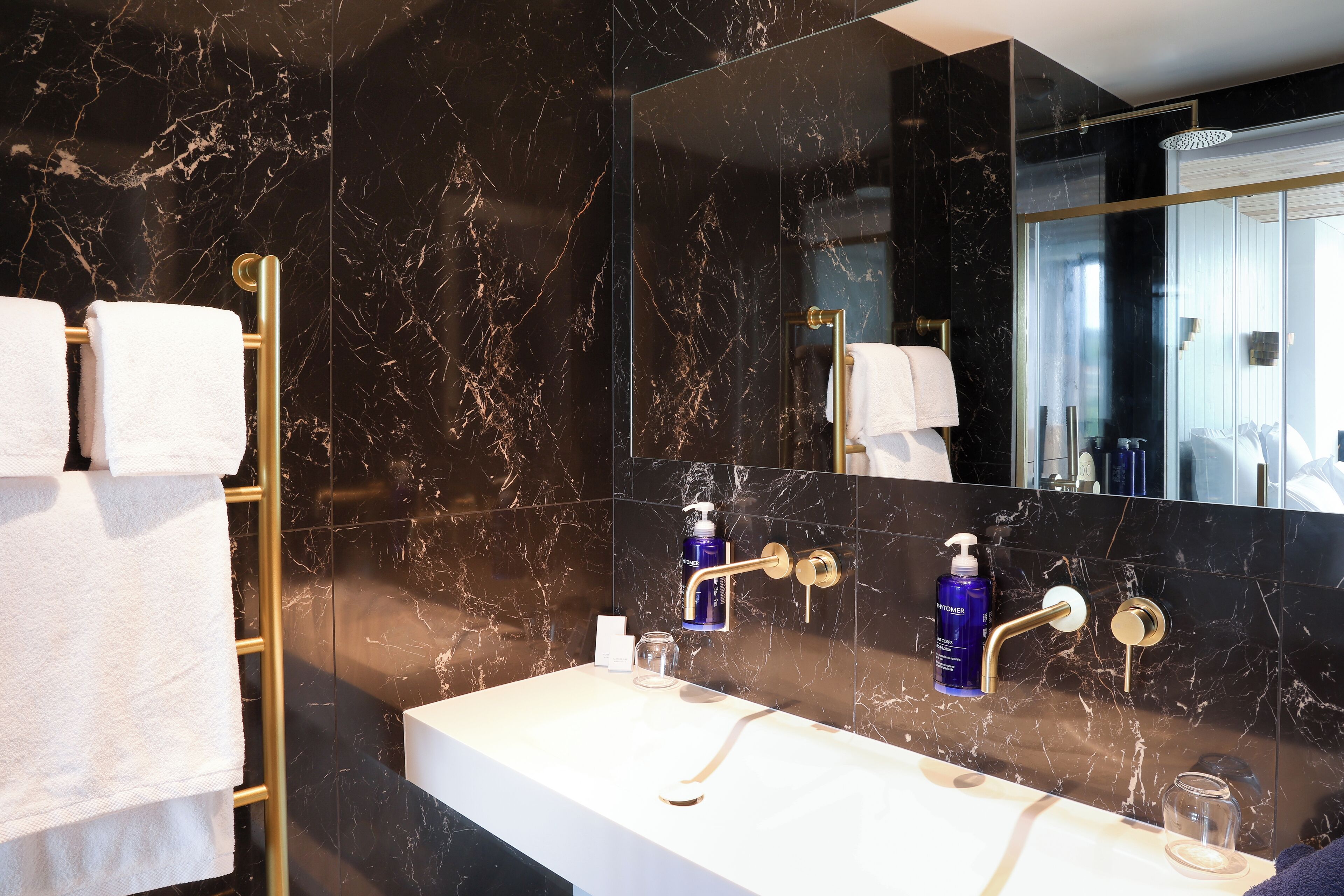 A modern bathroom with a marble backsplash, a gold towel rack, a white sink, and a large mirror.