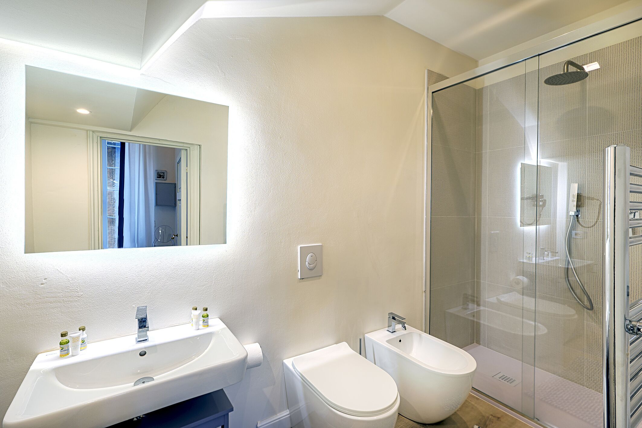 A modern bathroom with a large mirror, two sinks, a toilet, a bidet, and a glass-enclosed shower.