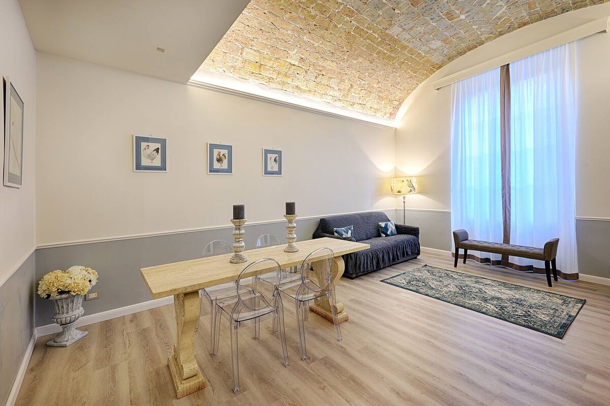 Ein modernes Wohnzimmer mit einem Holzesstisch, transparenten Stühlen, einer Couch und einer Bank. An der Wand hängen gerahmte Bilder, auf dem Tisch steht eine Vase mit Blumen und es gibt ein großes Fenster mit transparenten Vorhängen.