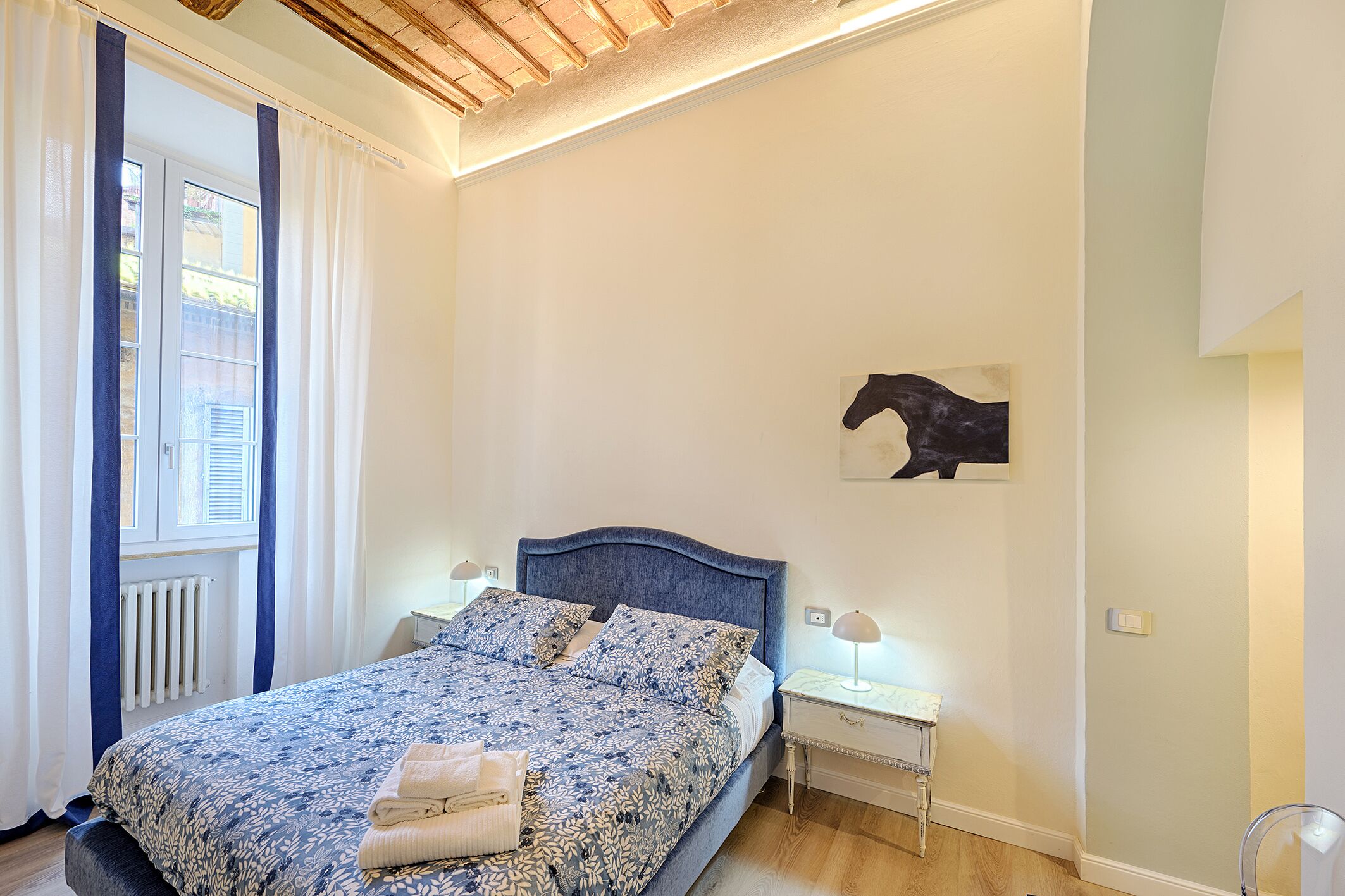 Un dormitorio con cama, un radiador, una ventana con cortinas, una obra de arte colgada en la pared y lámparas de noche.