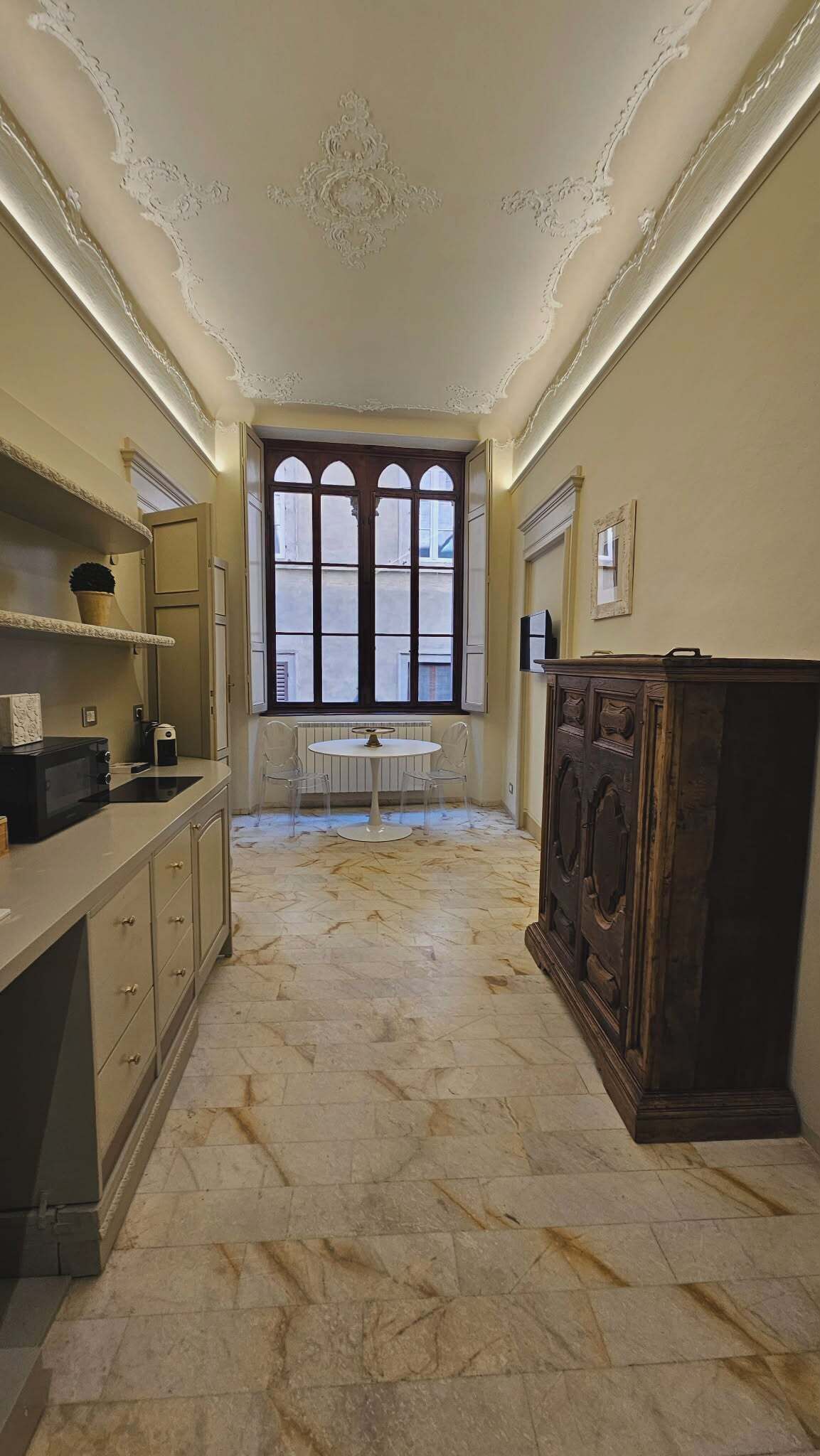 Una habitación con suelo de mármol, un ventanal con múltiples vidrios, una mesa redonda blanca con sillas y un mueble de madera oscura.