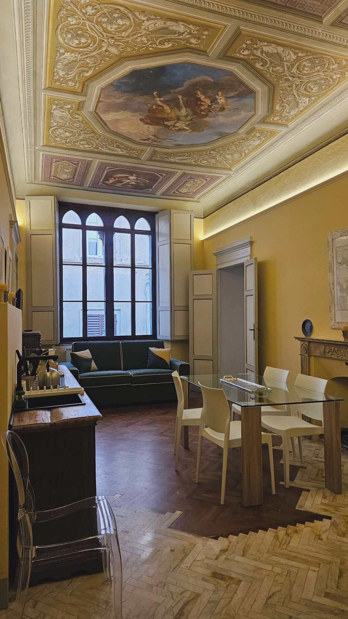 Een kamer met een groot raam, een eettafel met stoelen, een bank en een decoratief plafond.