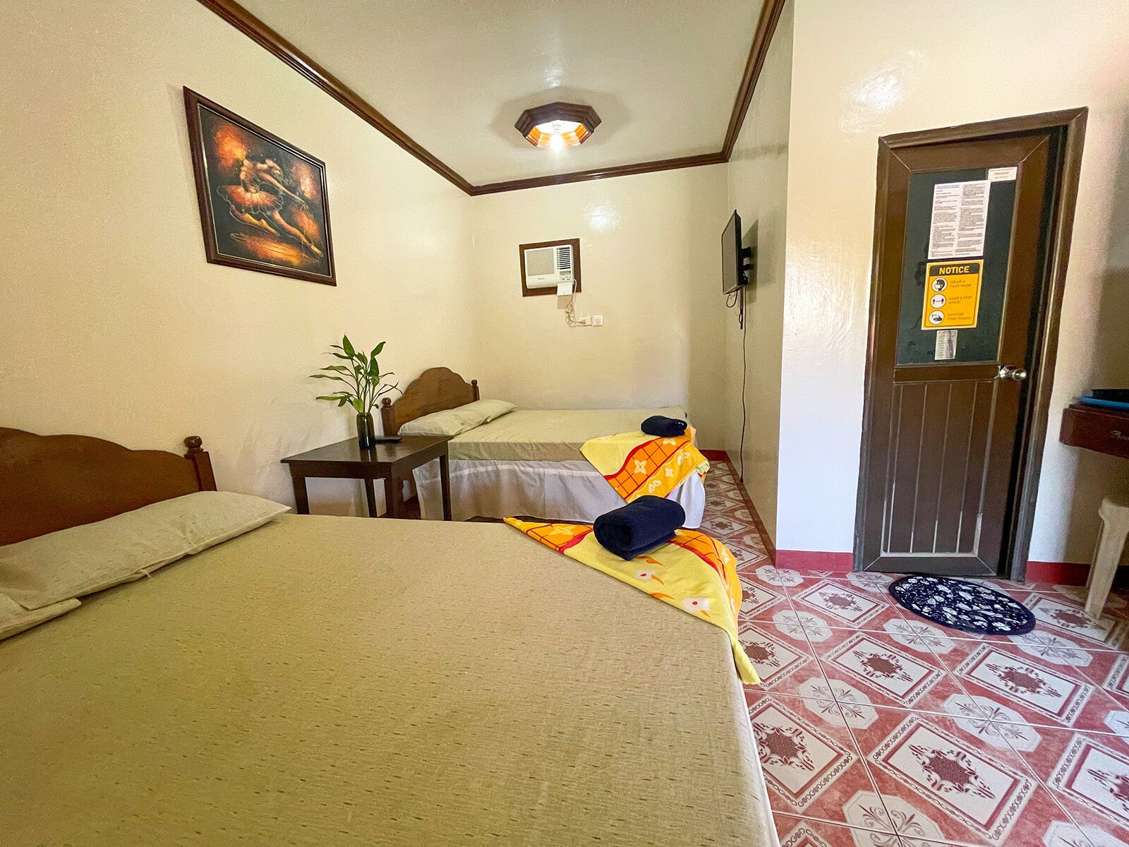 Une chambre d’hôtel avec deux lits, une petite table avec une plante, un tableau au mur, un avis sur la porte et des carreaux de sol à motifs.