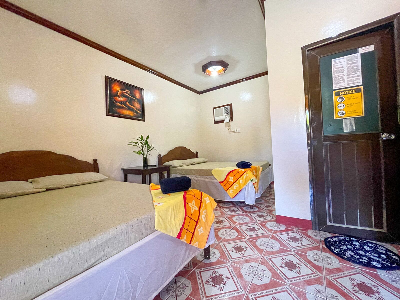 A hotel room with two beds, a small table with a plant, a wall-mounted air conditioner, and a door with a notice board.