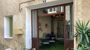 Une entrée d’immeuble avec une porte vitrée, un petit espace extérieur aménagé avec des chaises et un pot de fleurs.