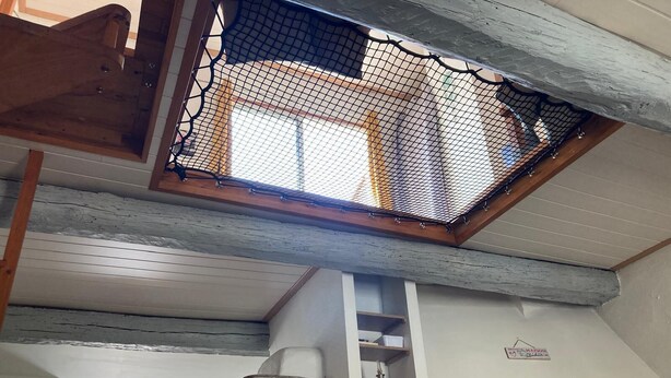 Une pièce avec un plafond en pente, une poutre en bois, un mur blanc orné d’un motif décoratif et une petite étagère sur laquelle sont posés quelques objets.