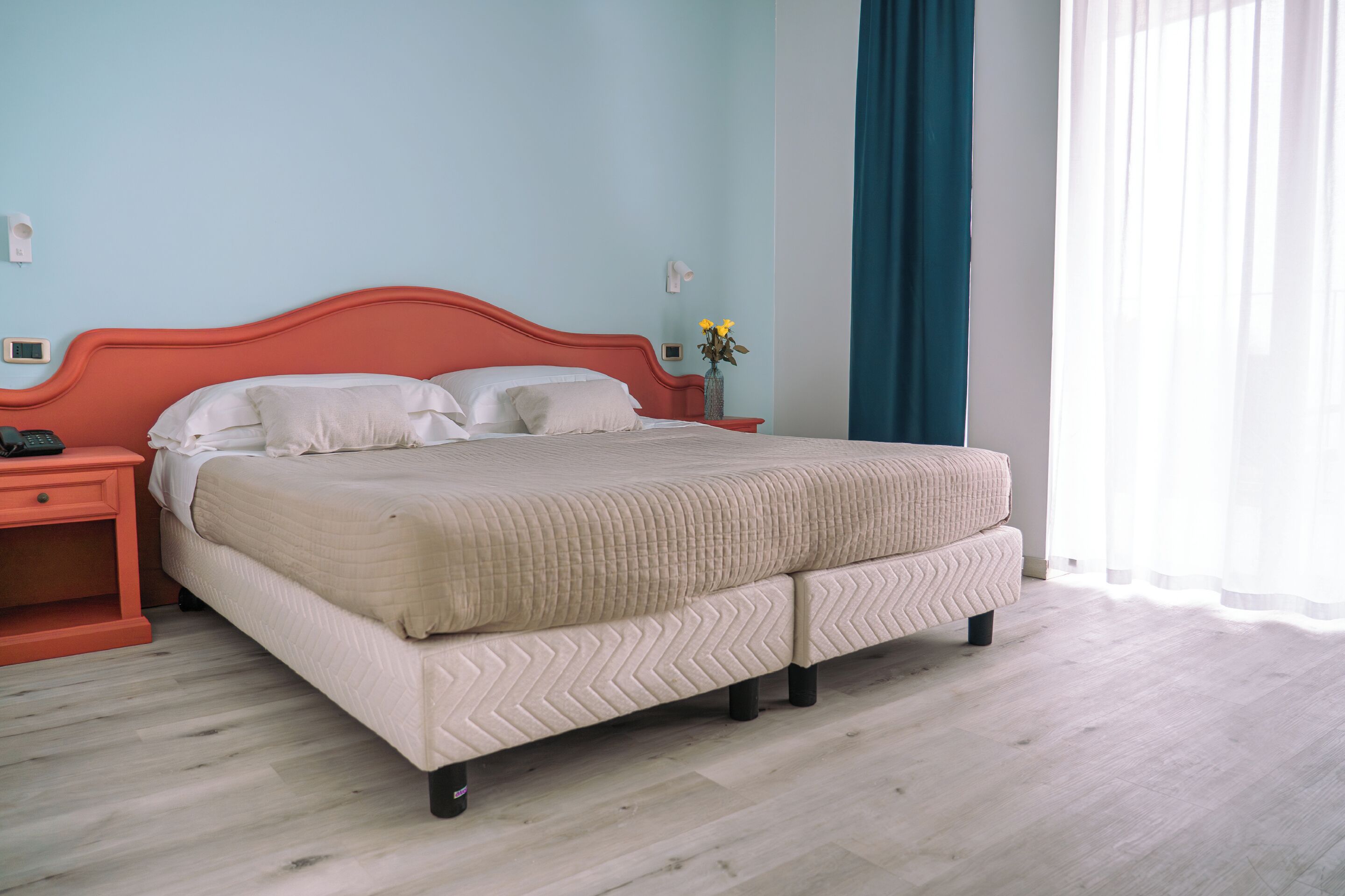 A neatly made bed with a quilted cover, a nightstand with a phone, and a vase with flowers on the wall.
