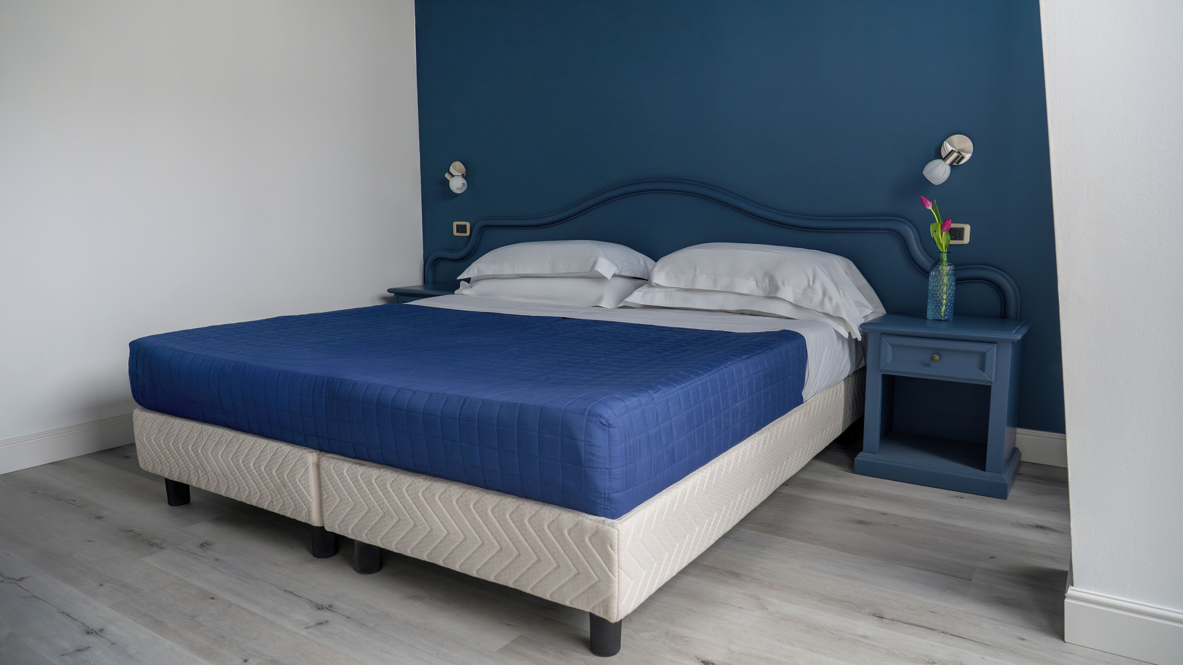 A bed with a blue bedspread, a headboard, and a nightstand with a vase.