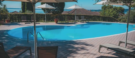 Una piscina con sillas de descanso y sombrillas, rodeada de un suelo de baldosas y follaje.