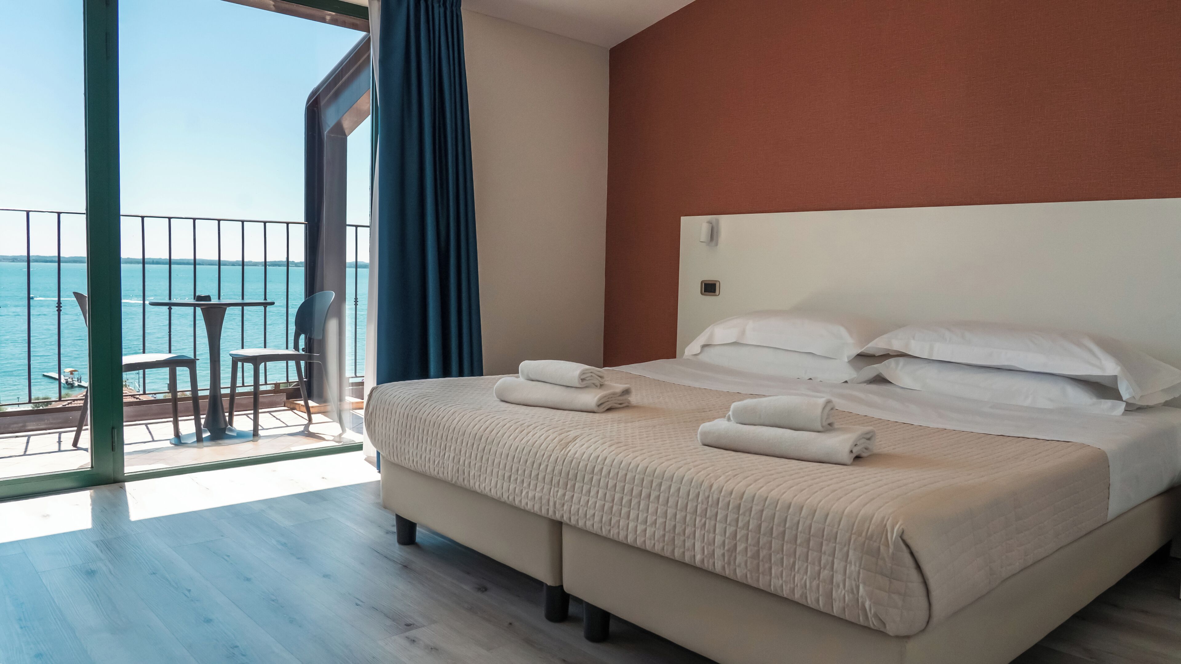 A hotel room with a large bed, two pillows, and two towels. There is a balcony with a table and chairs, and a view of the sea.