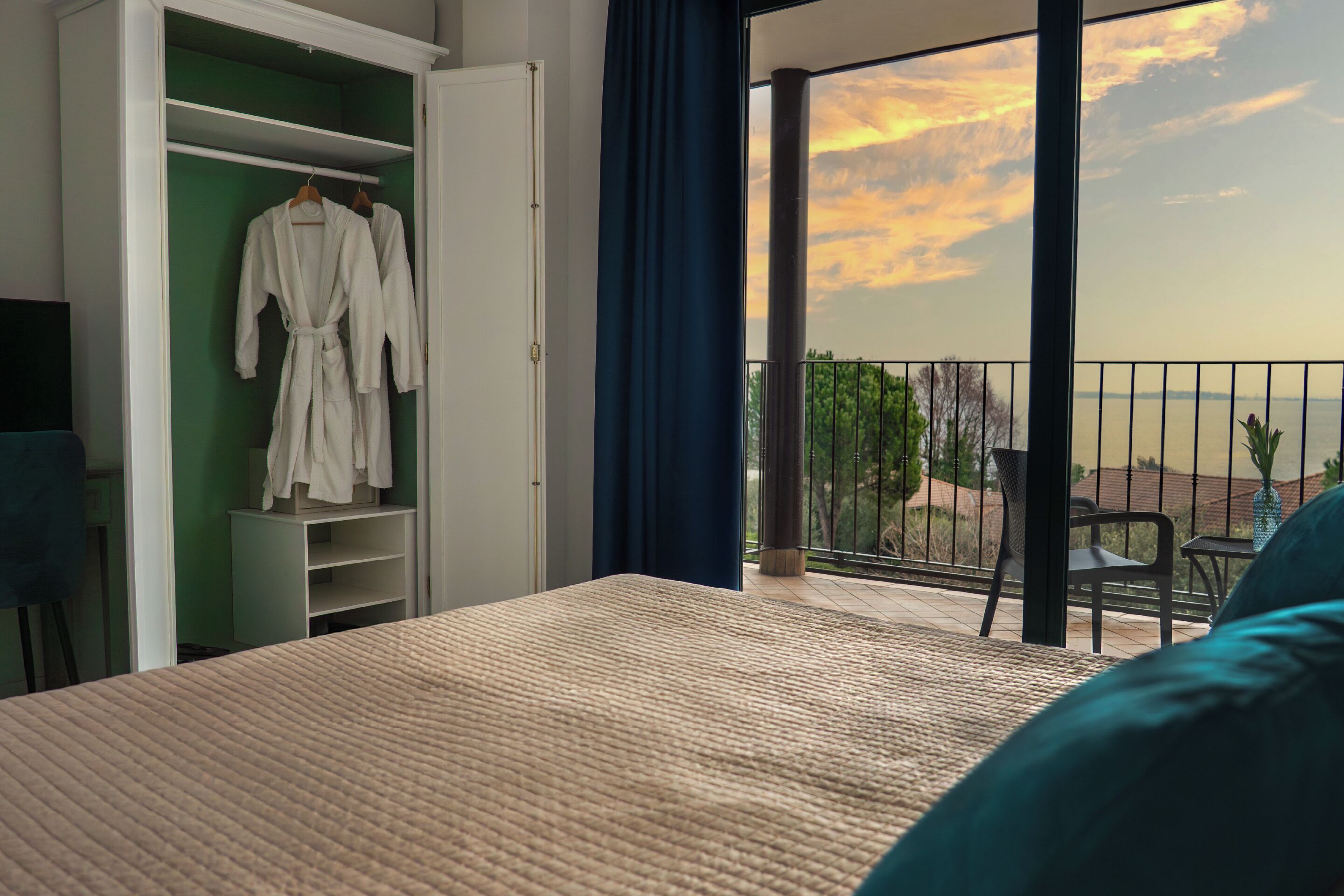 A bedroom with a bed, a wardrobe, and a balcony with a view of the sunset.