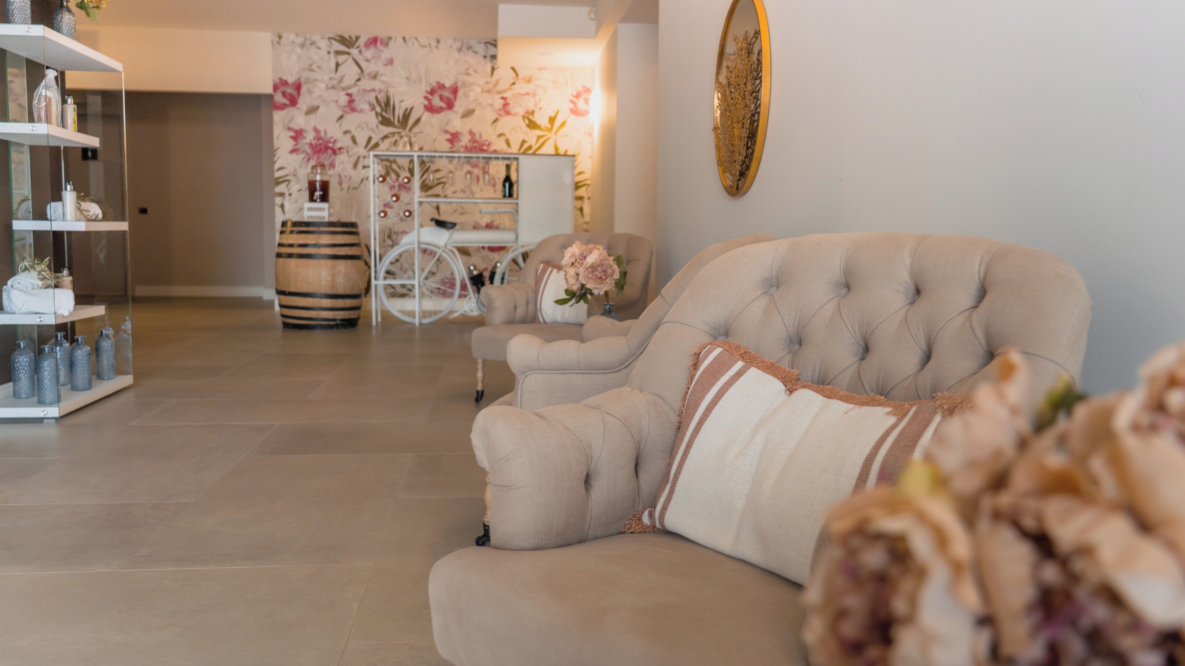 A living room with a beige sofa, a white coffee table, and a floral wallpaper. There is a white cabinet with glass doors on the left.