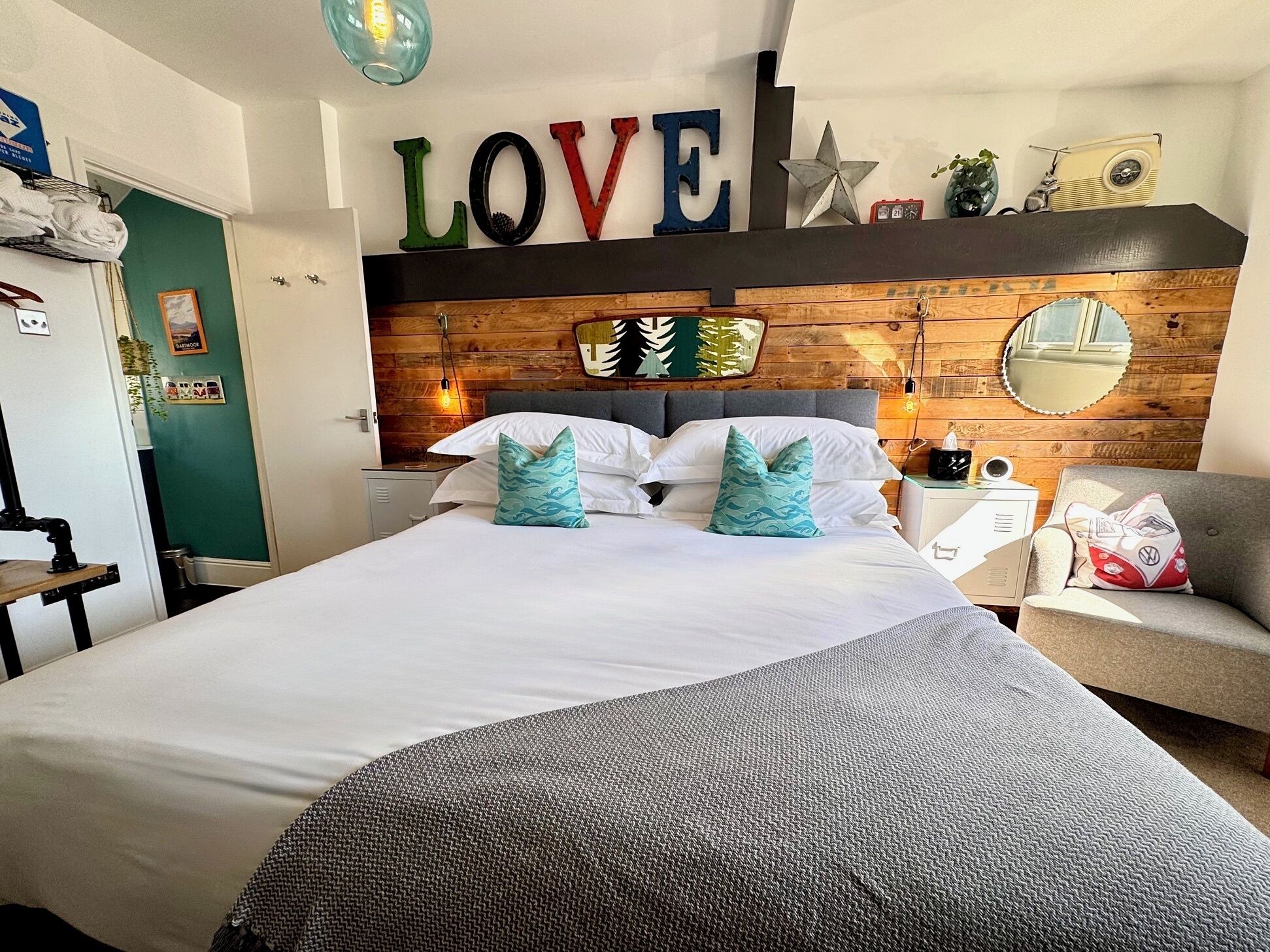 A neatly made bed with white linens and teal pillows, a wooden headboard with a 'LOVE' sign, and a vintage radio on a shelf.