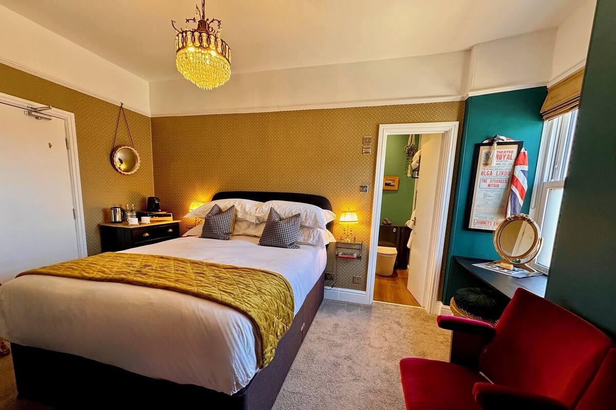 Een hotelkamer met een bed, een rode stoel, een kroonluchter, een spiegel en een klein bureau met een lamp.
