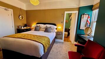 A hotel room with a bed, a red chair, a chandelier, a mirror, and a small desk with a lamp.
