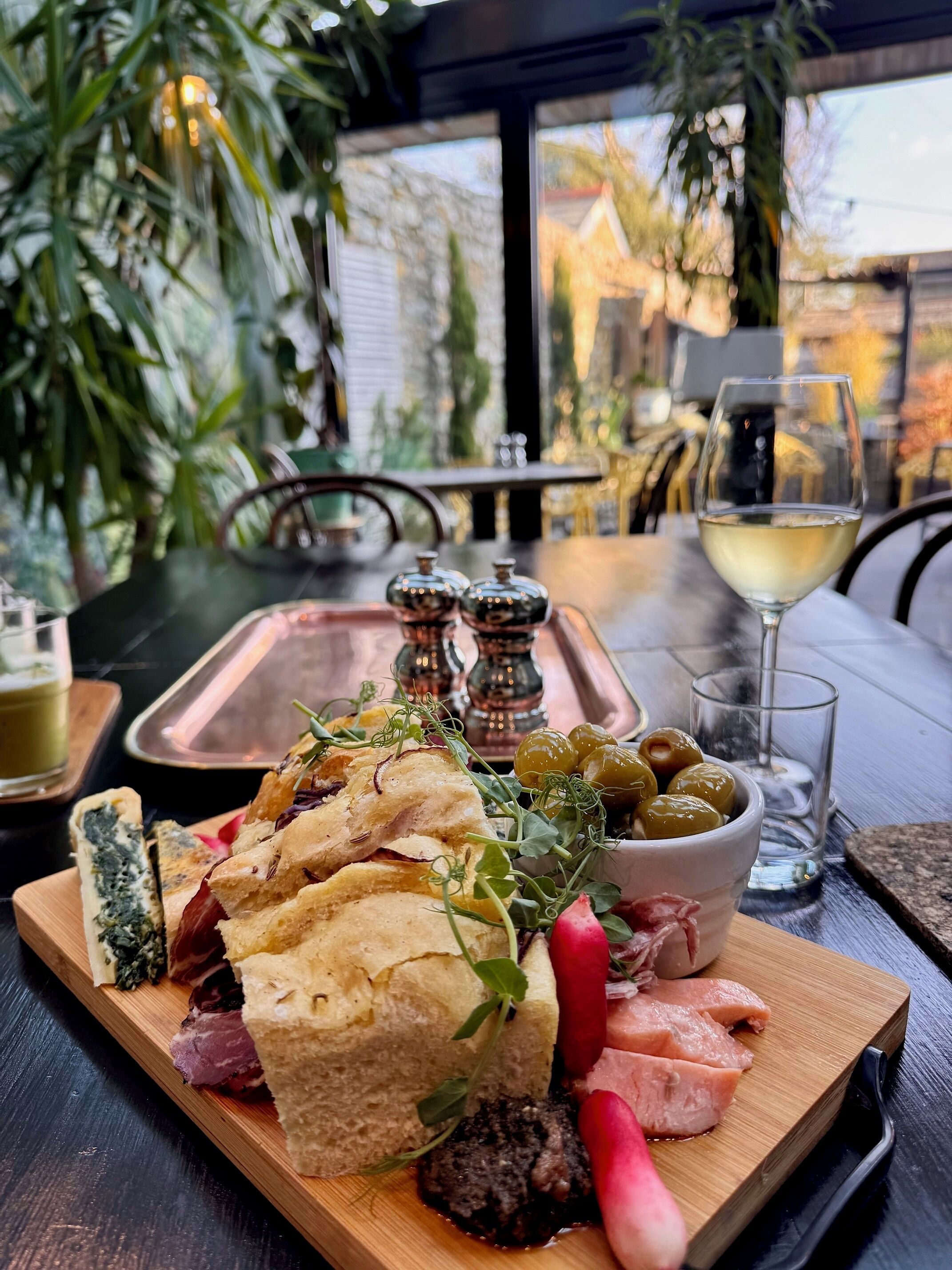 A wooden board with various foods including bread, cheese, olives, and meats, accompanied by a glass of white wine and a bowl of olives.
