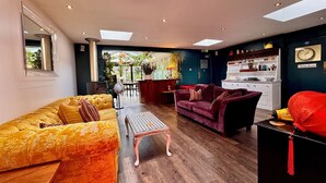 Un salon moderne avec un canapé jaune, un canapé violet et une table basse. On y trouve également un coin cuisine avec une horloge au mur.