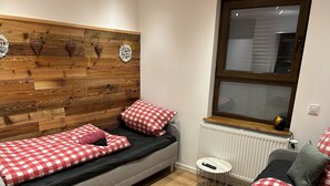 Una habitación pequeña y acogedora con cabecera de madera, una ventana con persianas, un radiador y una mesita redonda con un control remoto.
