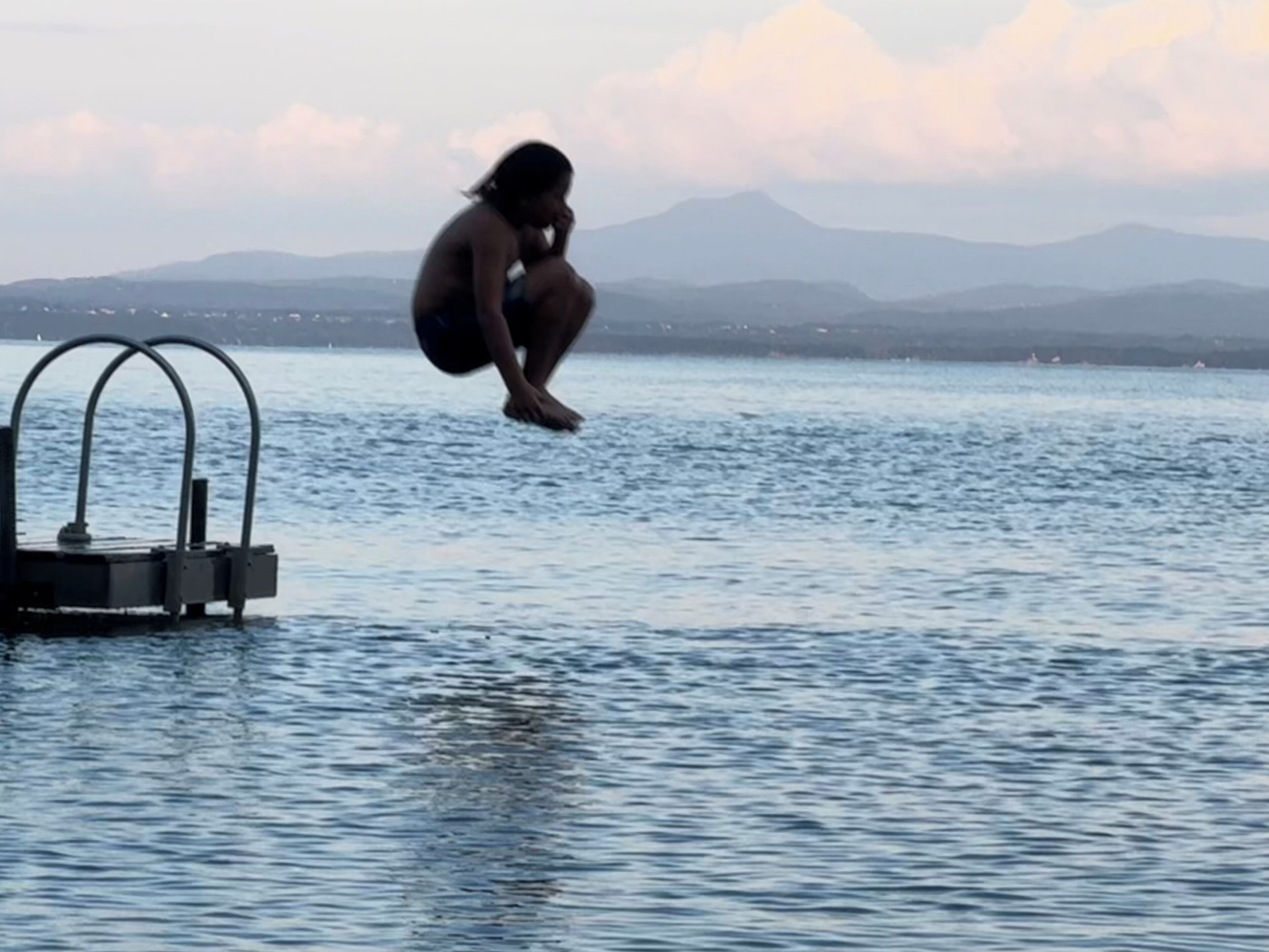 A person jumping off a diving board into a body of water with mountains in the background.
