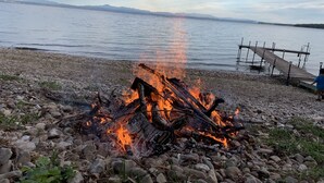 Vuur op een kiezelstrand met een houten steiger die zich uitstrekt over kalm water, omgeven door bergen.