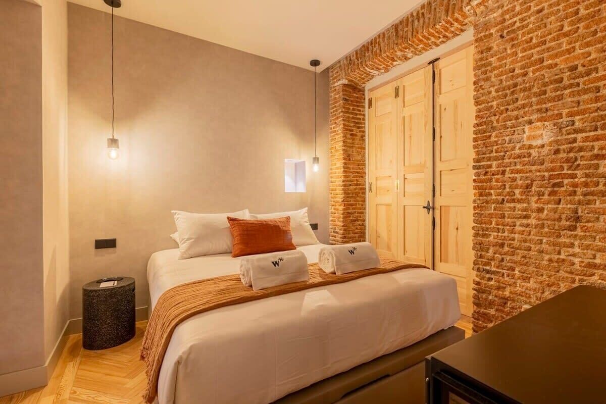 Une chambre d’hôtel avec un lit, un banc en bois, une poubelle, une lampe fixée au mur et un mur en briques.