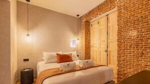 Une chambre d’hôtel avec un lit, un banc en bois, une poubelle, une lampe fixée au mur et un mur en briques.