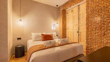 Une chambre d’hôtel avec un lit, un banc en bois, une poubelle, une lampe fixée au mur et un mur en briques.