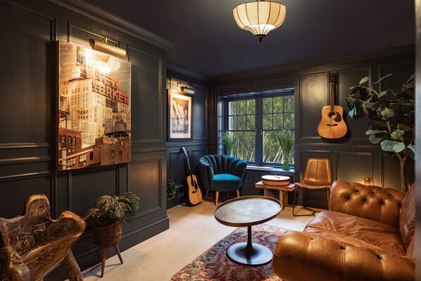 A room with a dark color scheme, featuring a leather sofa, a round table, a guitar, and a large window with a view of greenery.