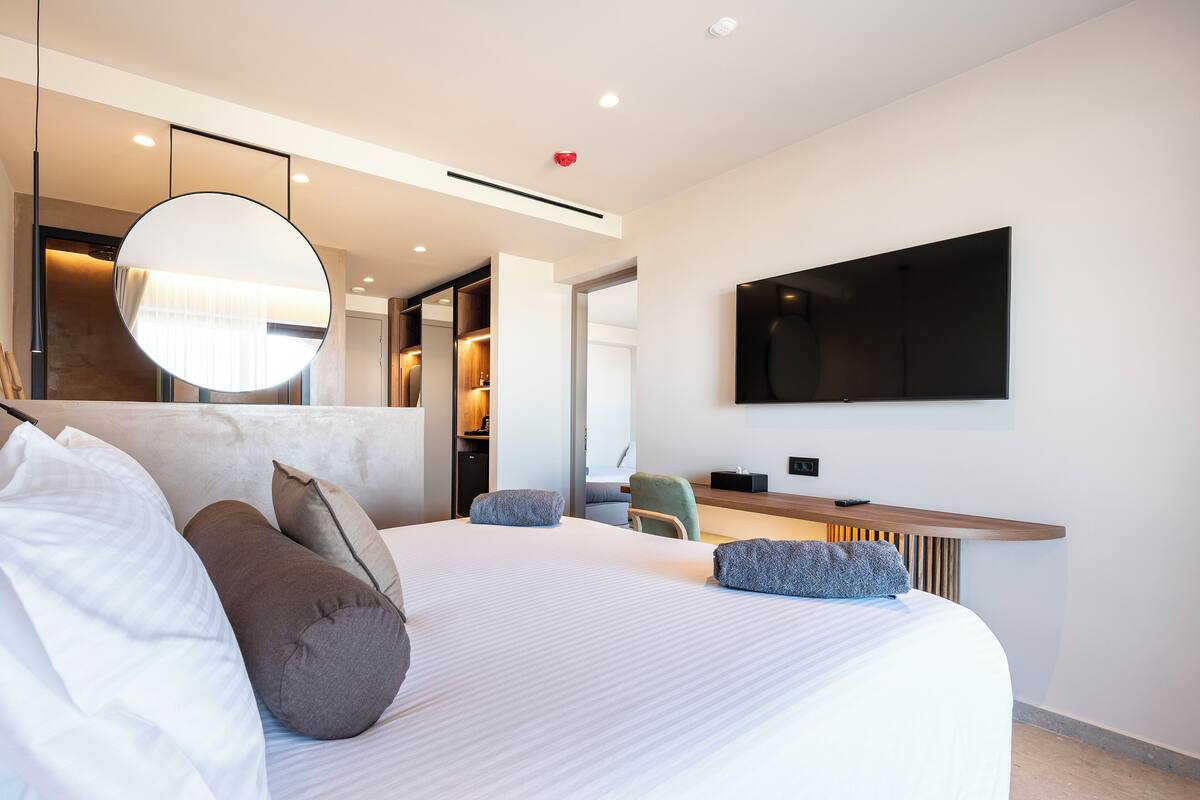 A modern hotel room with a large bed, a TV mounted on the wall, and a mirror above the bed.