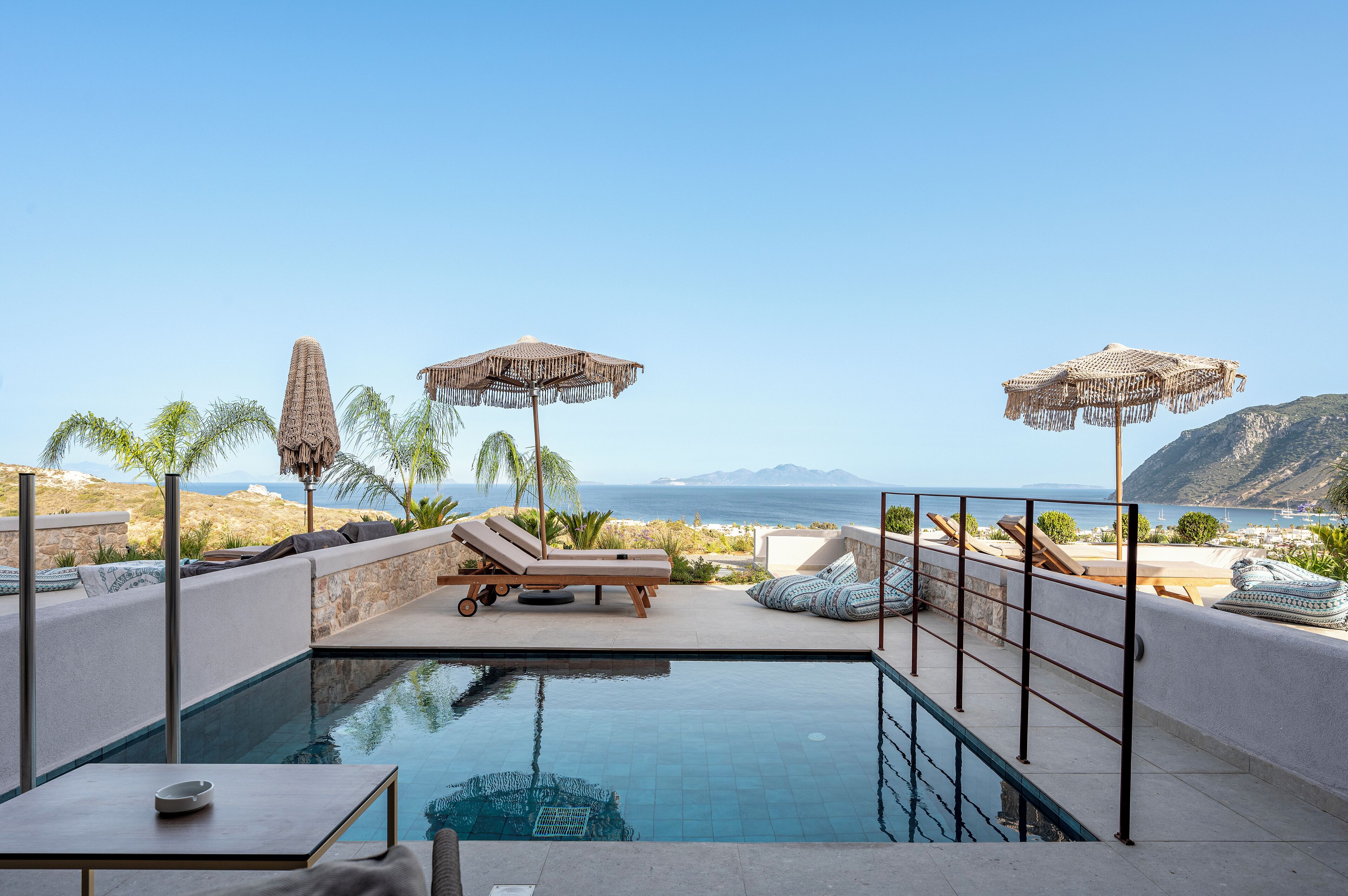 A poolside area with lounge chairs and umbrellas, overlooking a beach and mountains.