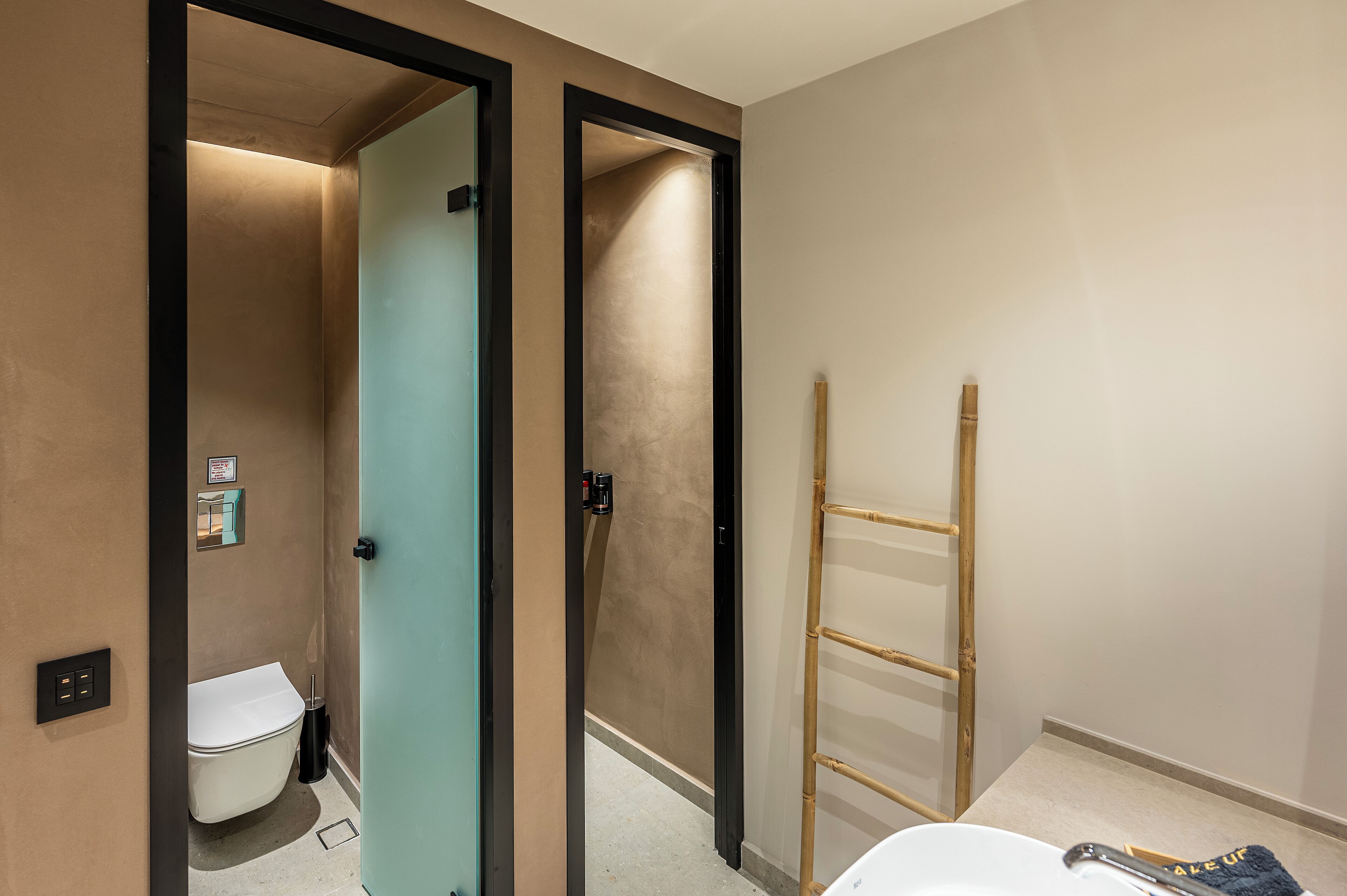 Une salle de bain comprenant des toilettes, un lavabo et un espace douche. Une échelle en bambou est appuyée contre le mur.