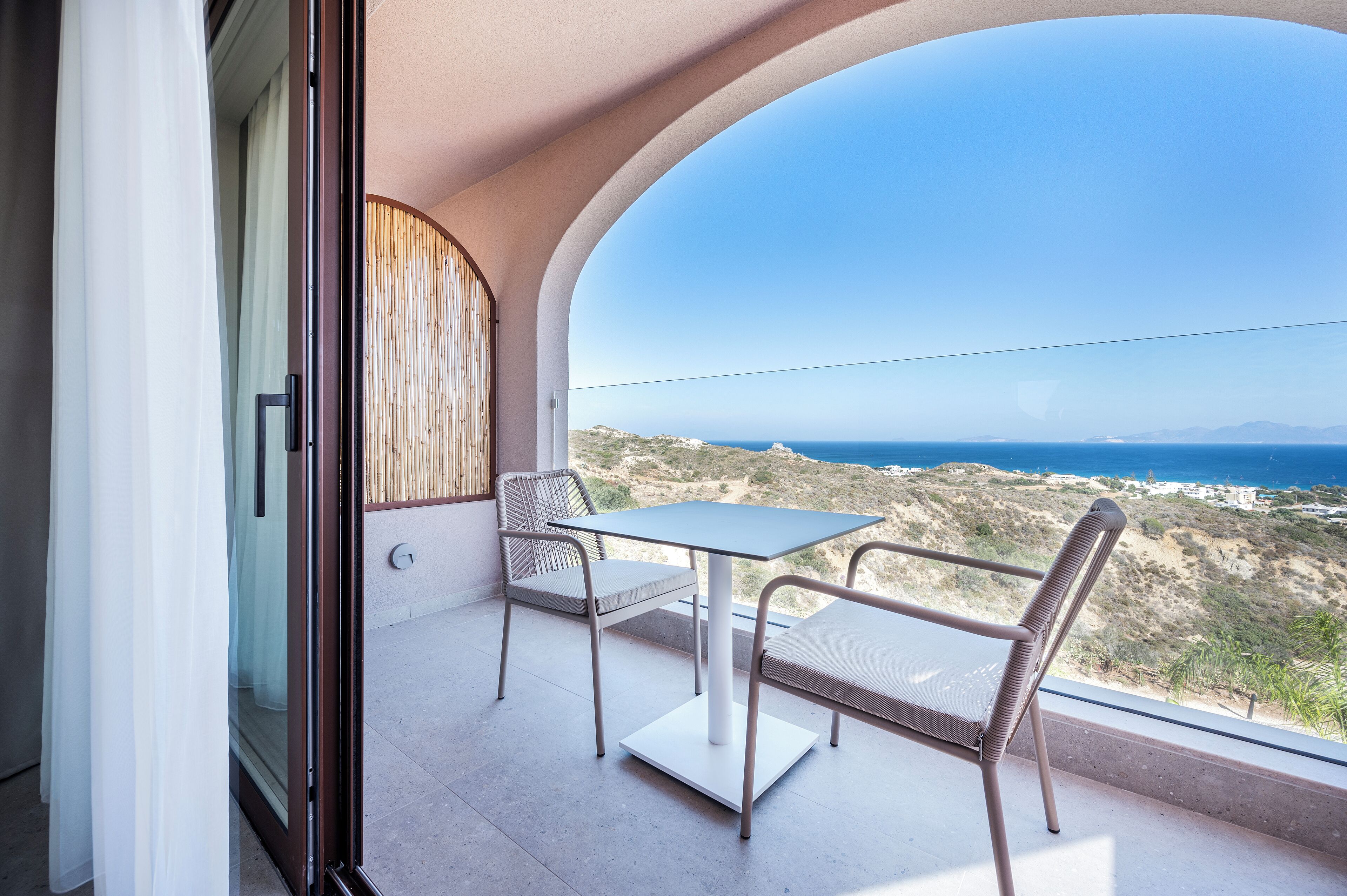 A balcony with a table and chairs, offering a view of the coastline and mountains.