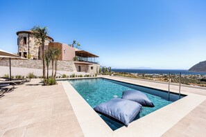 A modern pool area with clear blue water, two blue lounge chairs, and a view of the sea and mountains.