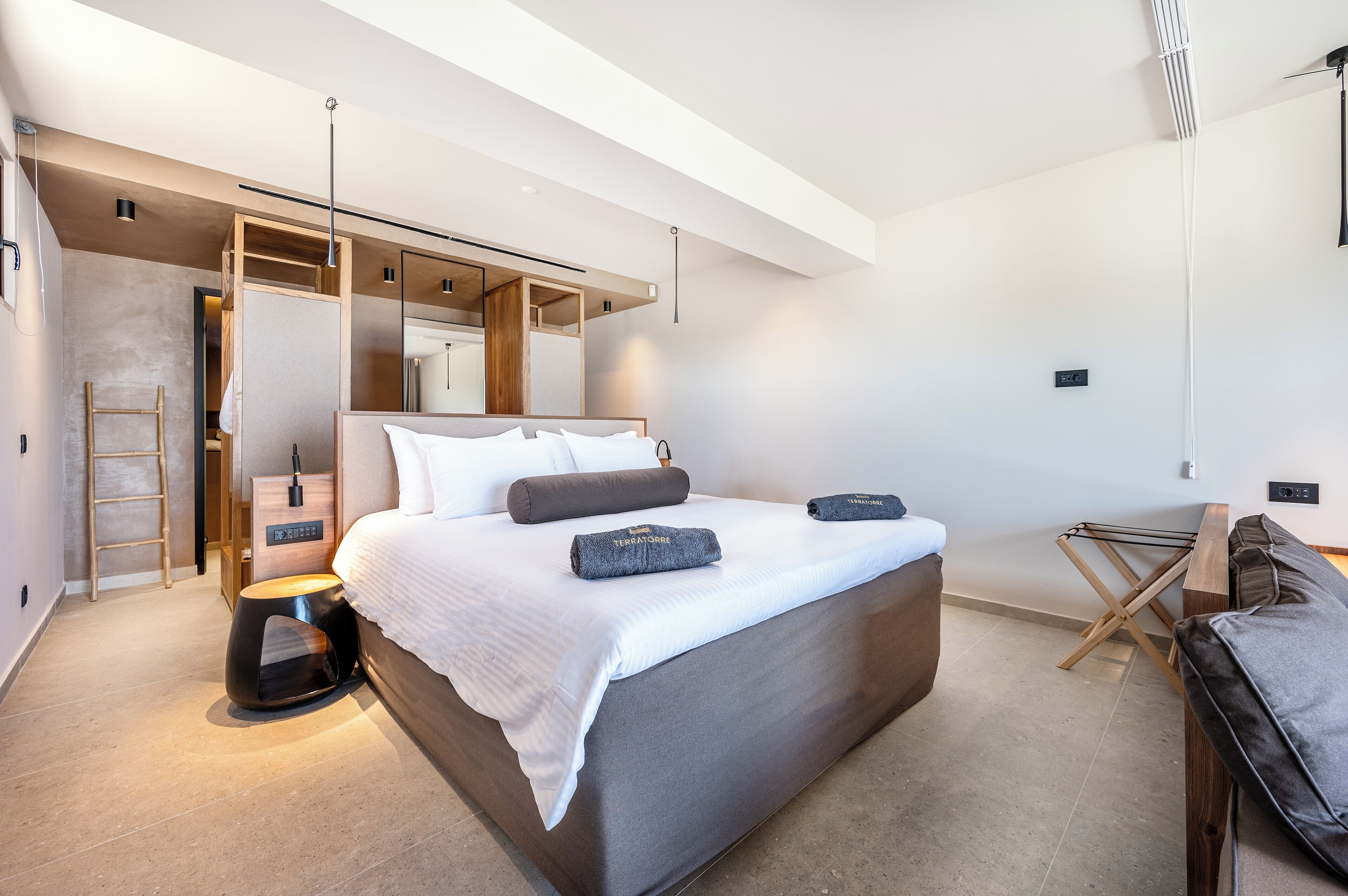 A modern bedroom with a large bed, a wooden ladder, a small stool, and a hanging light fixture.
