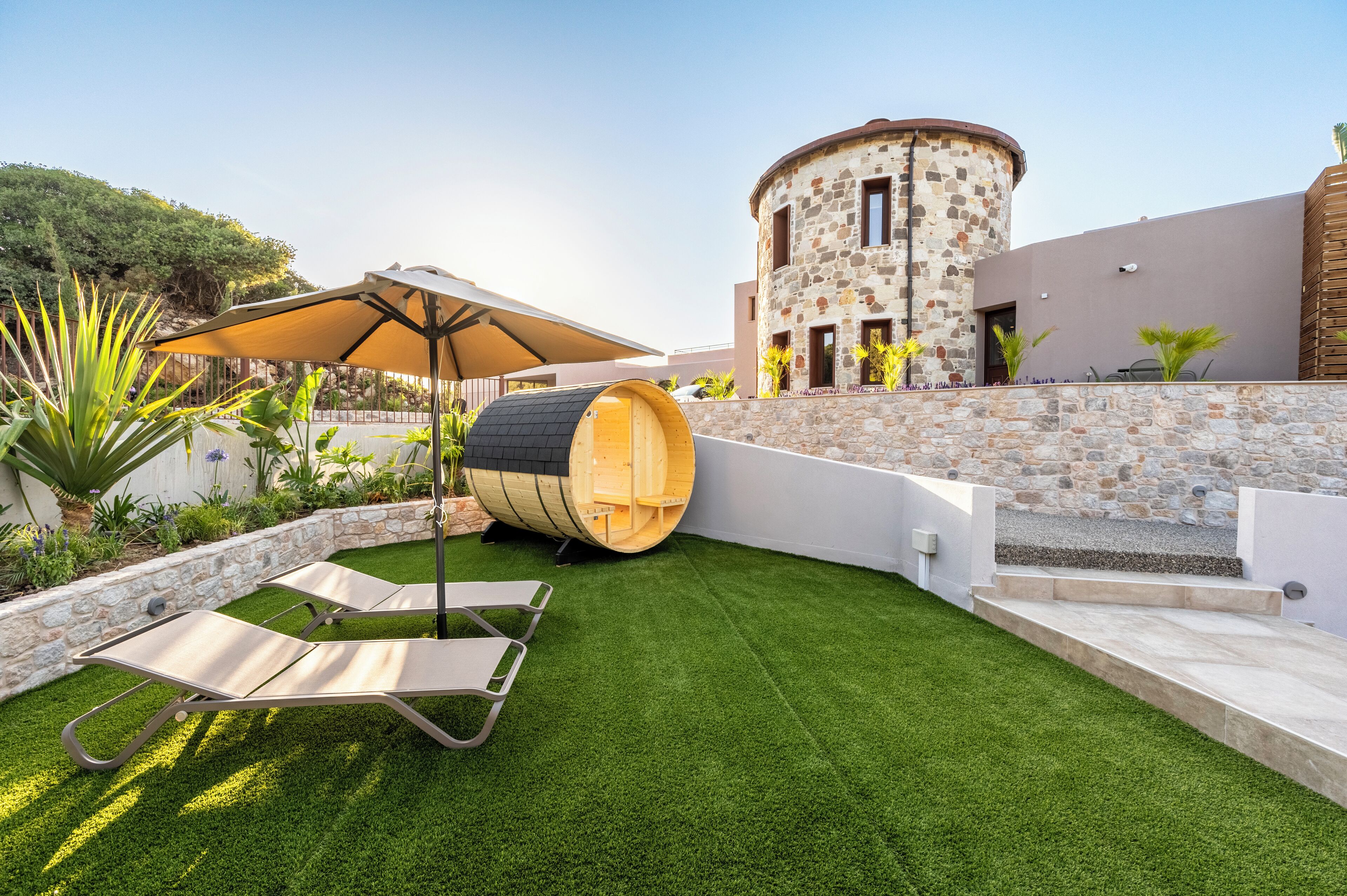 Un espace extérieur moderne avec une pelouse artificielle, un tonneau en bois et un bâtiment en pierre doté d’une tour cylindrique.
