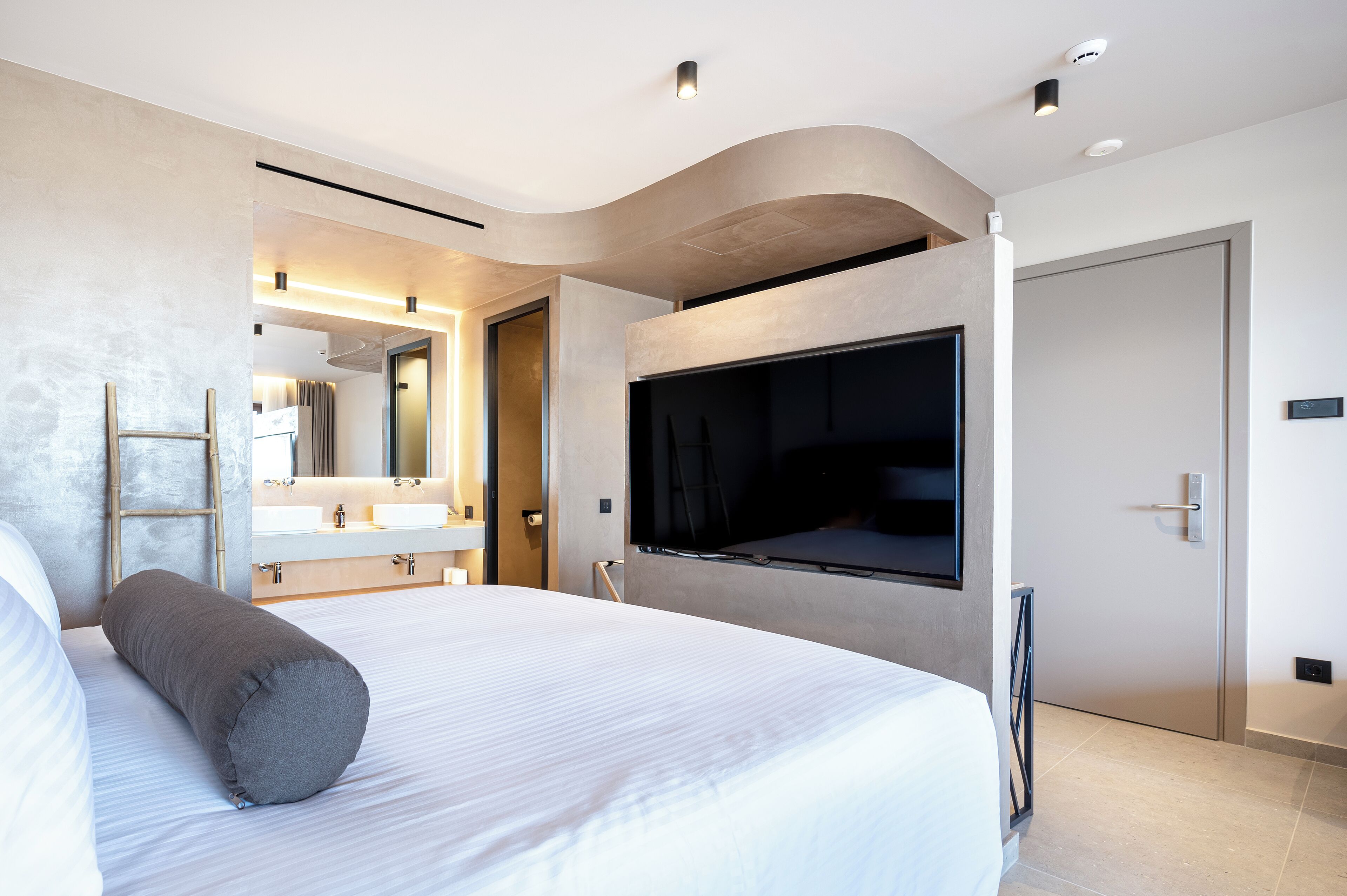 A modern hotel room with a large bed, a TV mounted on the wall, a bathroom with a vanity, and a door with a handle.