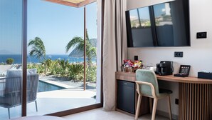 A modern hotel room with a large TV, a desk with coffee maker and snacks, a chair, and a view of the ocean and palm trees. - TERRATORRE (Kos)