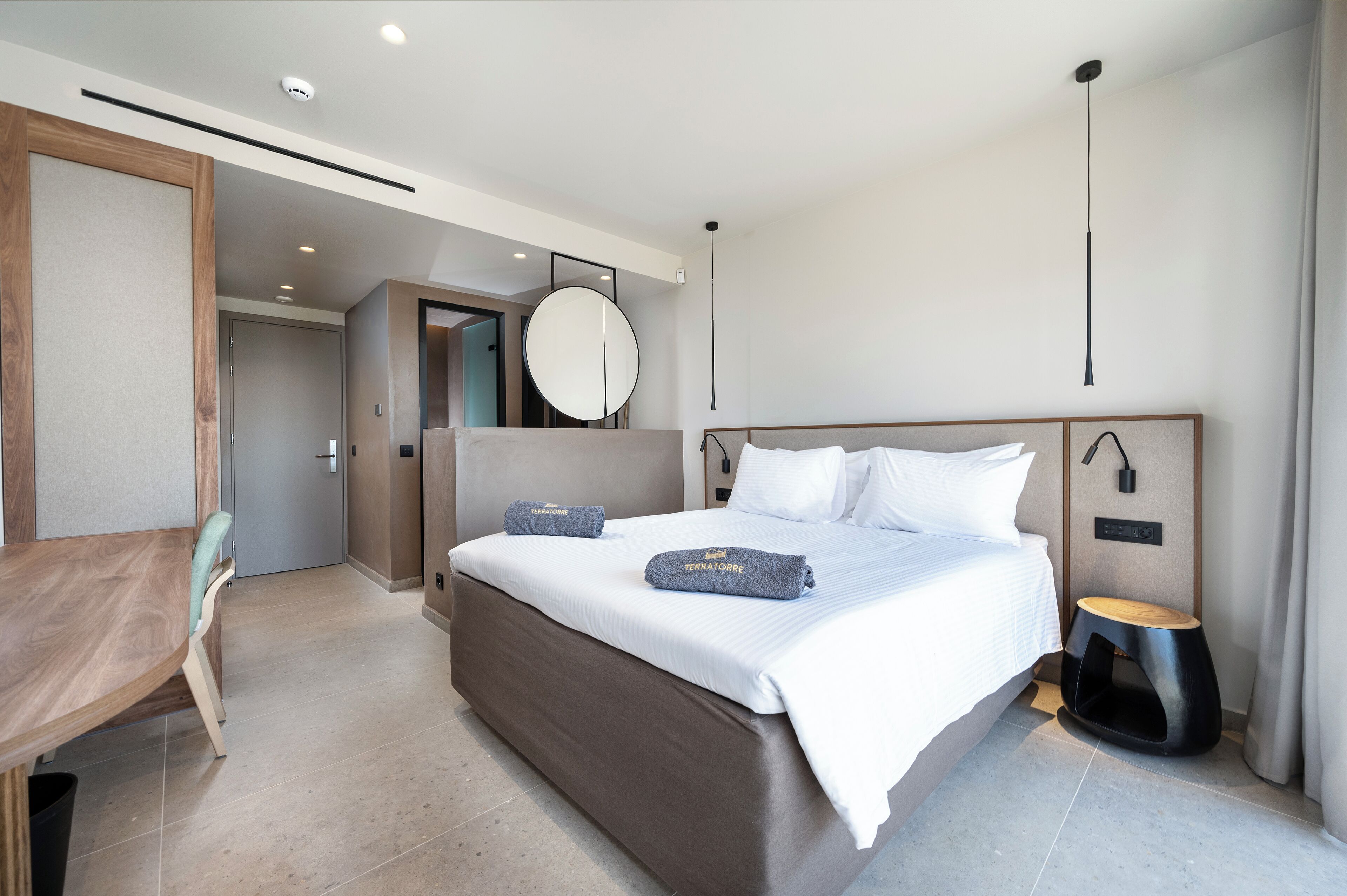 A modern hotel room with a large bed, bedside tables, a round mirror, and a wooden desk.