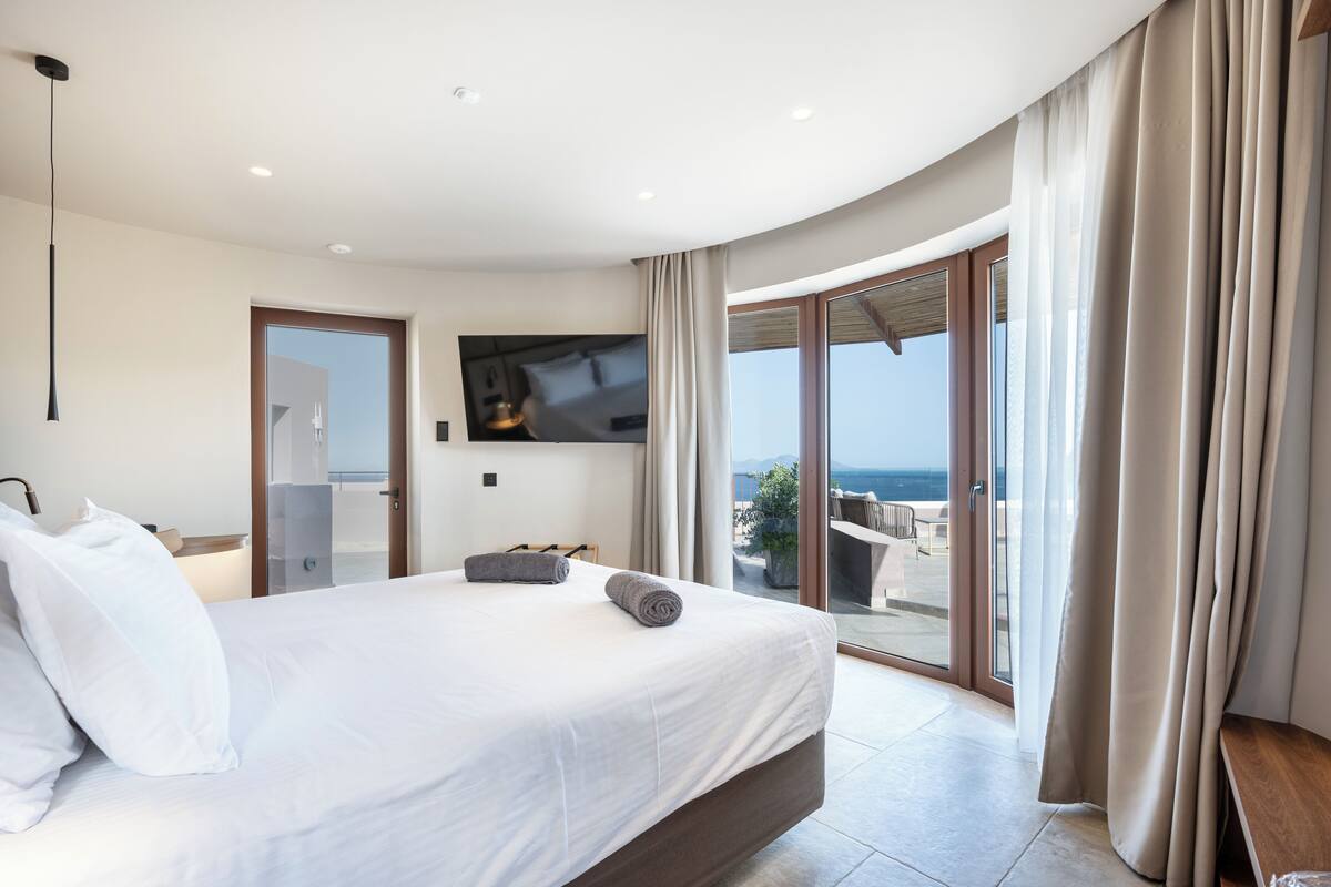 A modern hotel room with a large bed, a flat-screen TV, and a balcony with a view of the sea.