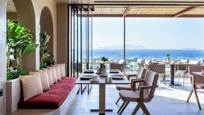 A modern dining area with a view of the sea, featuring a long table, cushioned seating, and potted plants. - TERRATORRE (Kos)