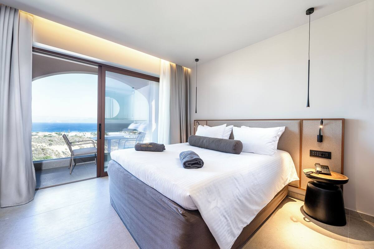 A modern hotel room with a large bed, a bedside table with a phone, and a view of the sea.