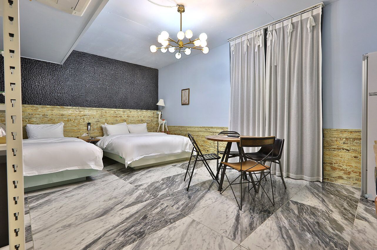 Habitación de hotel con dos camas, una mesa de comedor con sillas, un candelabro y un ventanal con cortinas.