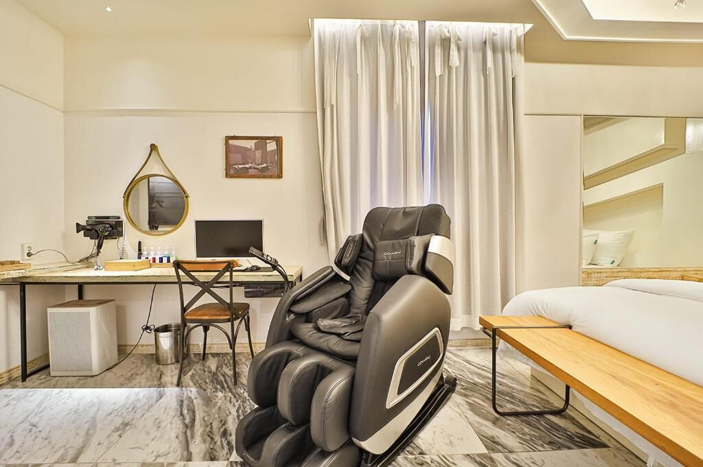 Habitación de hotel con sillón de masaje, escritorio con computadora, cama y ventana grande con cortinas.
