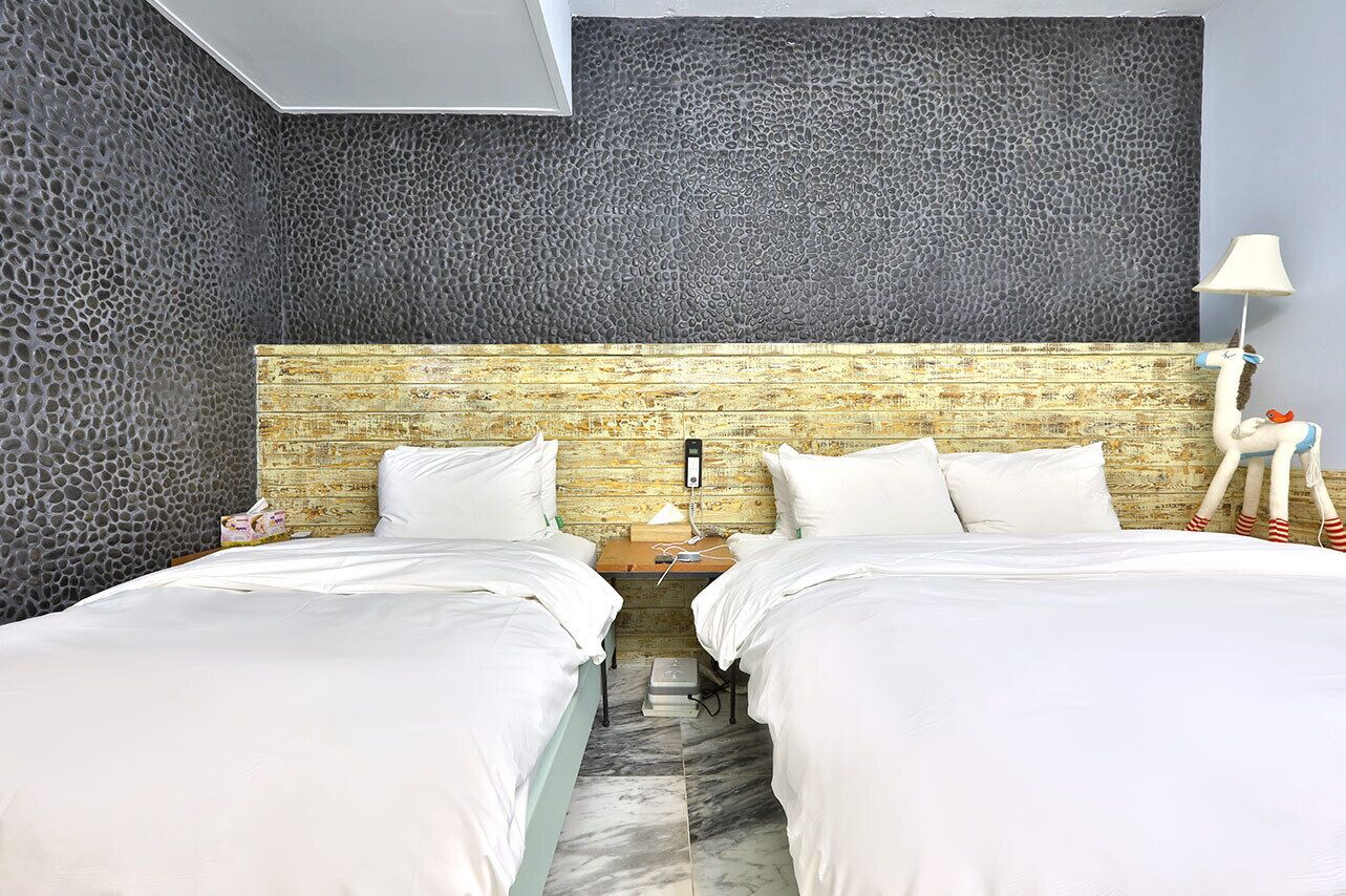 A hotel room with two beds, a textured wall, a wooden headboard, a nightstand with a lamp, and a suitcase.