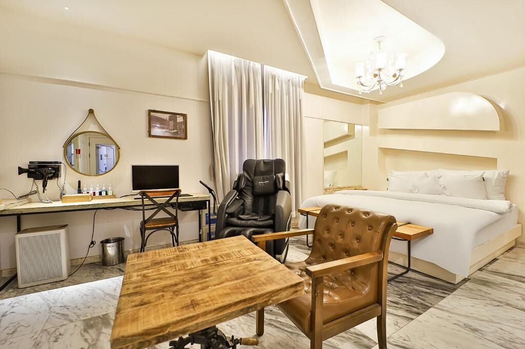 A hotel room with a bed, a desk with a computer, a massage chair, a wooden table, and a chandelier.
