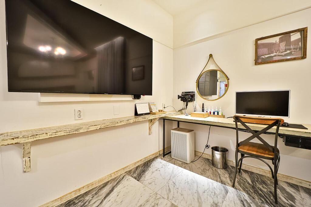 A hotel room with a large flat-screen TV mounted on the wall, a small desk with a chair, a mirror, and a radiator.