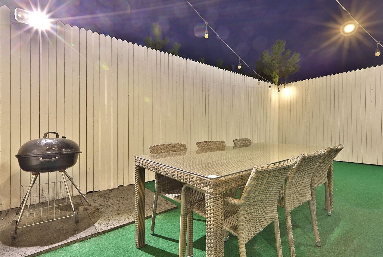An outdoor patio with a glass-top table and wicker chairs, a barbecue grill, and string lights against a white fence.